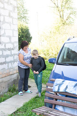 A grandmother walks a young boy, wearing a backpack, to a car. She touches his shoulder with one...