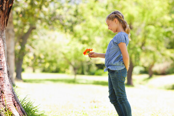 Girl holding orange daisy-like flower near large tree trunk in sunlit grassy park, gazing at petals