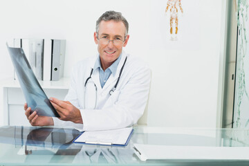 Adult male doctor holding x-ray film with stethoscope while sitting at glass-top desk in exam room