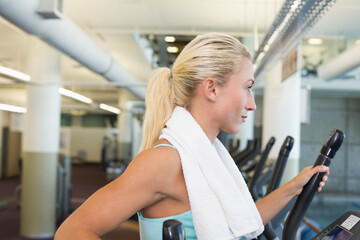 Woman in turquoise tank top wearing gym towel exercising on elliptical machine by ducts, copy space