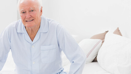 Senior man sitting on bed wearing blue striped pajamas and smiling in bedroom with white linens