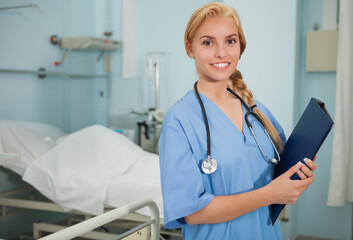 Female nurse standing next to bed in patient room holding clipboard and stethoscope, copy space
