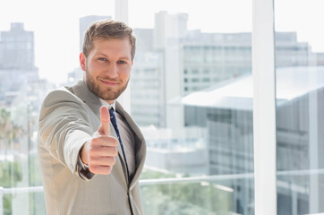Businessman in suit giving thumbs-up standing in office with railing viewing cityscape, copy space