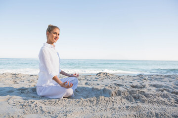 Woman meditating cross-legged on sandy shoreline facing calm sea wearing white clothing, copy space