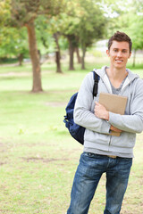 Male student standing in park holding hardcover notebook and carrying navy backpack, copy space