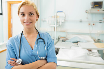 Adult female nurse wearing scrubs and stethoscope standing by bed in hospital room, copy space