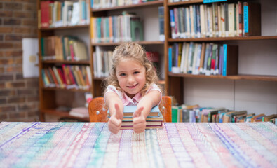 Female child student leaning on stacked books on patterned tablecloth in library giving thumbs up
