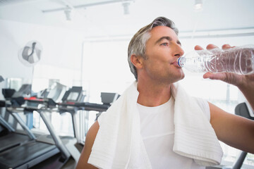 Adult male drinking water from clear plastic water bottle at gym with towel, treadmills, copy space