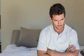 Man wearing white polo sitting on bed edge gazing in bedroom with taupe pillows, copy space