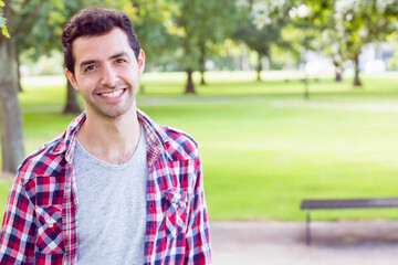 Standing in urban park wearing plaid shirt by wooden bench and paved path, copy space