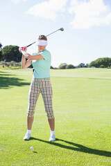 Golfer gripping club, aiming swing at golf ball teed on sunlit fairway bordered by leafy trees