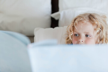Child boy reading pale blue hardcover book while lying on bed among white pillows, copy space