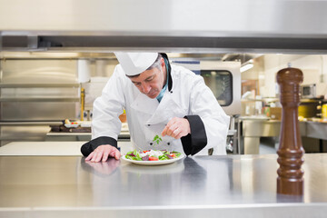 Male chef adding fresh herbs to salad on plate beside wooden pepper mill in professional kitchen