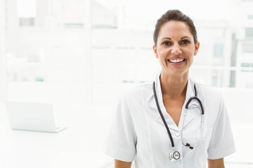 Female doctor wearing white uniform and stethoscope standing by windows with laptop, copy space