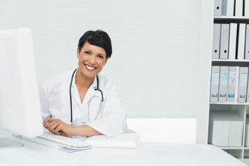 Female physician sitting at desk in office using monitor while wearing lab coat with stethoscope