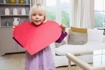 White female child standing by grey shelving unit in living room holding large red paper heart