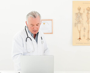 Male doctor wearing white coat using laptop in office with stethoscope, certificate, skeleton chart