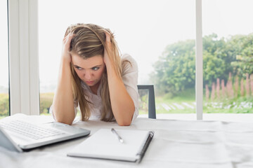 Woman holding head, gazing at desk with laptop, notebook and pen by window with garden view