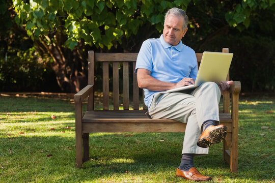 Senior man in polo shirt and khaki trousers sitting on wooden bench in garden using laptop
