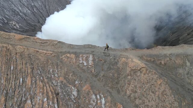 Aerial view of woman walking along the rim of steaming Mount Bromo volcano crater in Indonesia. Concept of nature, travel, adventure, and breathtaking landscapes.