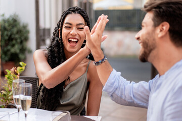Happy diverse couple celebrating success with a high five, laughing and drinking champagne outdoors.