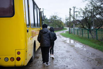 Two people walking toward a yellow bus on a wet road in a rural setting, obscured by an overcast sky, creating a somber and melancholic mood.
