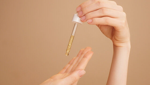 Close-up of female hands on isolated beige background holding dropper, anti-aging facial serum, booster dripping on palm.Commercial skincare, moisturizing, vitamin c, anti-wrinkle products advertising
