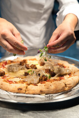 Professional chef garnishing a gourmet pizza with fresh microgreens, featuring unique toppings like fish, eggplant, and cheese, in a bustling restaurant kitchen