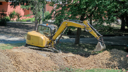 Large excavator digs a trench in the courtyard of a multi-story building to lay pipes.