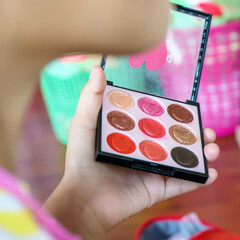 A hand holding a small square makeup palette with 9 vibrant cream shades in red, pink, brown, and peach tones.
