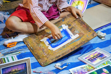 A child seated on the ground using pastel or crayon to create a blue artwork on a taped canvas board.