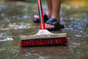 A person's hand holding an old long-handled scrub brush is scrubbing the cement floor, which is covered with algae stains.
