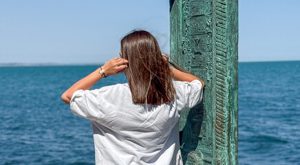 A young woman of undefined ethnicity stands thoughtfully by the water, adjusting her hair against a...