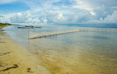 A serene coastal scene with calm, shallow waters, traditional fishing boats, and net fences...