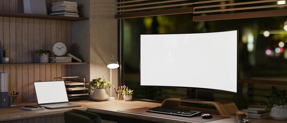 Blank curved screen computer and laptop under lamp light on wooden table in dim lit room or office.