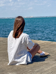 A young woman with medium-length brown hair sits cross-legged on a pier, gazing at the serene ocean...