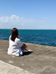 A young woman with long dark hair sits on a dock, gazing peacefully at the tranquil blue waters...