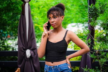 A serious young african woman stands with one hand on her hip while talking on her phone in an...