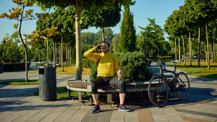 Elderly man in cycling gear drinking from thermos while sitting next to bike. Concept of healthy...