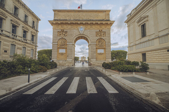 View of the historic Porte du Peyrou archway stands proudly, framed by symmetrical buildings and a zebra crossing, under a sky with scattered clouds, Montpellier, Occitanie, France.