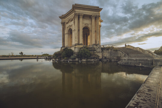 View of the serene water reflecting the architectural beauty of a historic structure under a dramatic sky, stairs leading up, Montpellier, Occitanie, France.