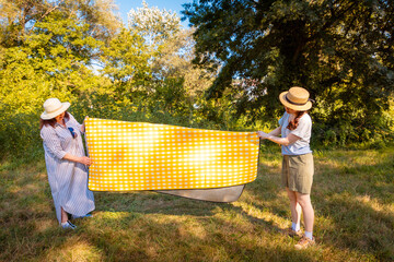 Caucasian mom and her young daughter unfold yellow picnic mat in green city park on sunny day....