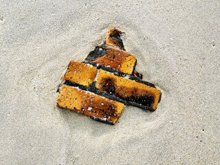 A piece of burnt and broken brick lies partially buried in fine beach sand, blending elements of...