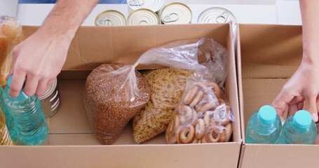Volunteer hands put canned food, cereals, butter into boxes, top view, close-up. Preparatory work for the distribution of aid to those in need. Sorting food products at a charity foundation warehouse - Powered by Adobe