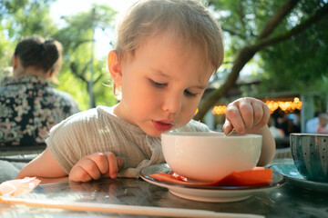 Cute little child enjoying healthy soup during family lunch at cozy street cafe. Authentic...