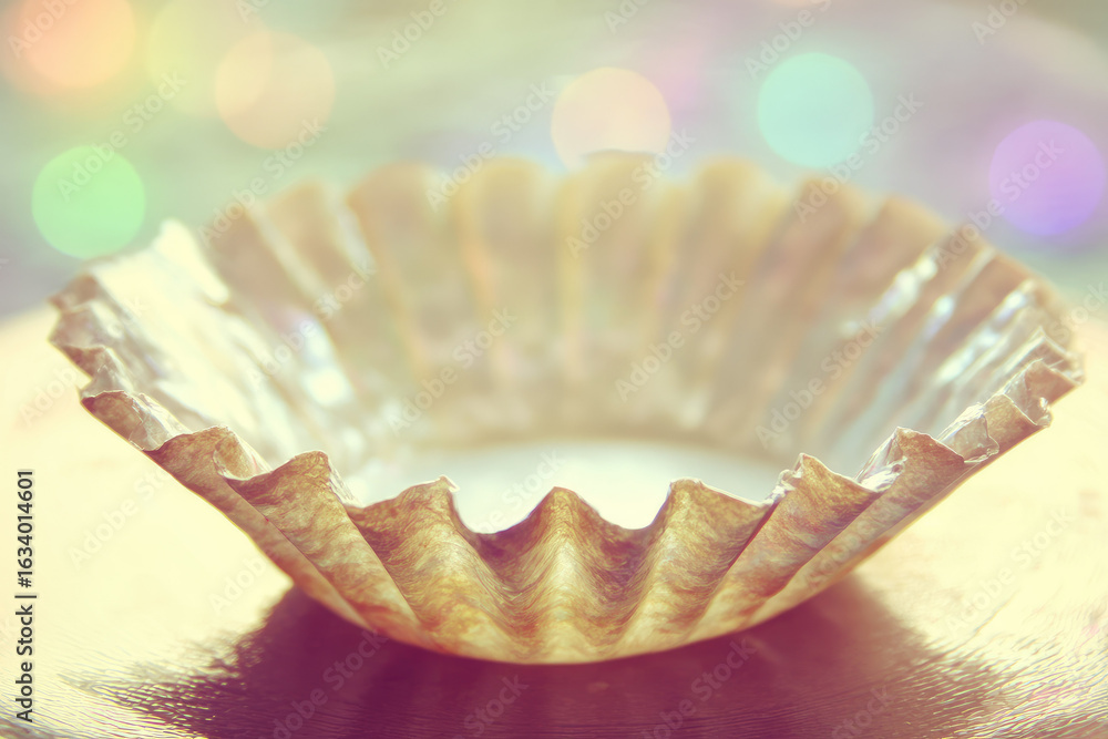 Poster empty cupcake wrapper with metallic sheen sits flattened on soft plate, surrounded by colorful bokeh