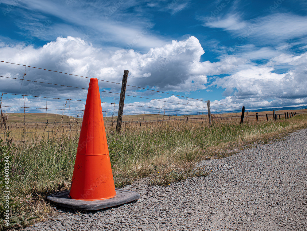 Aufkleber orange traffic cone on a gravel path in front of a fence, with grassland and clouds in the backgroun