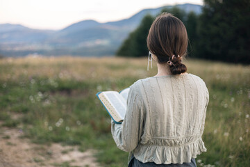 Young woman holding Bible in field with mountain view, copy space.