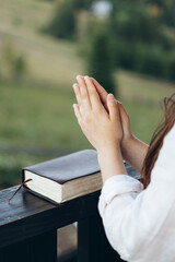 Woman praying with Bible on balcony overlooking peaceful mountain landscape.