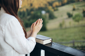 Woman praying with Bible on balcony overlooking peaceful mountain landscape.